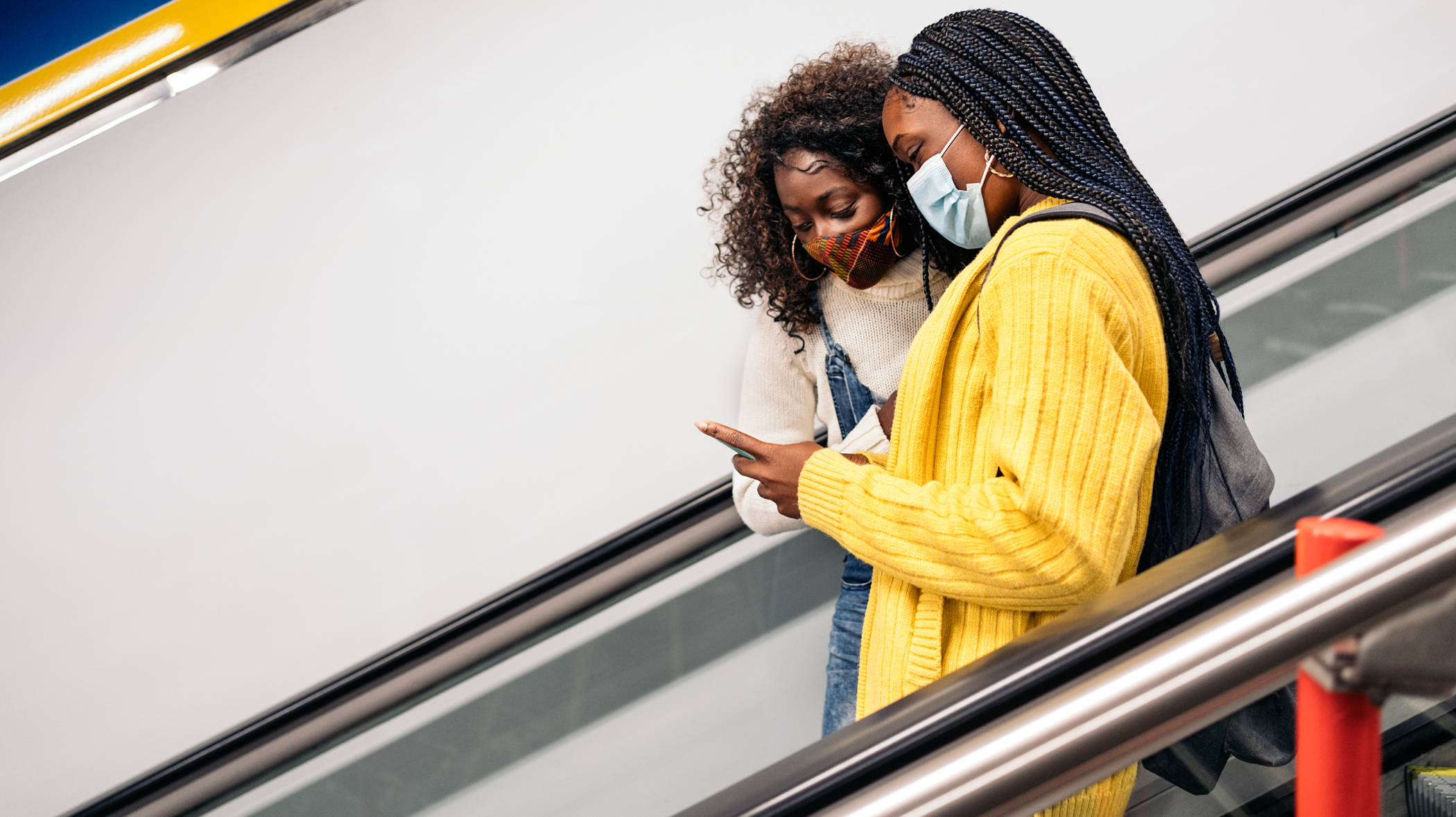 Stock photo of young Black friends wearing face mask due to covid19 looking at phone while using escalators.